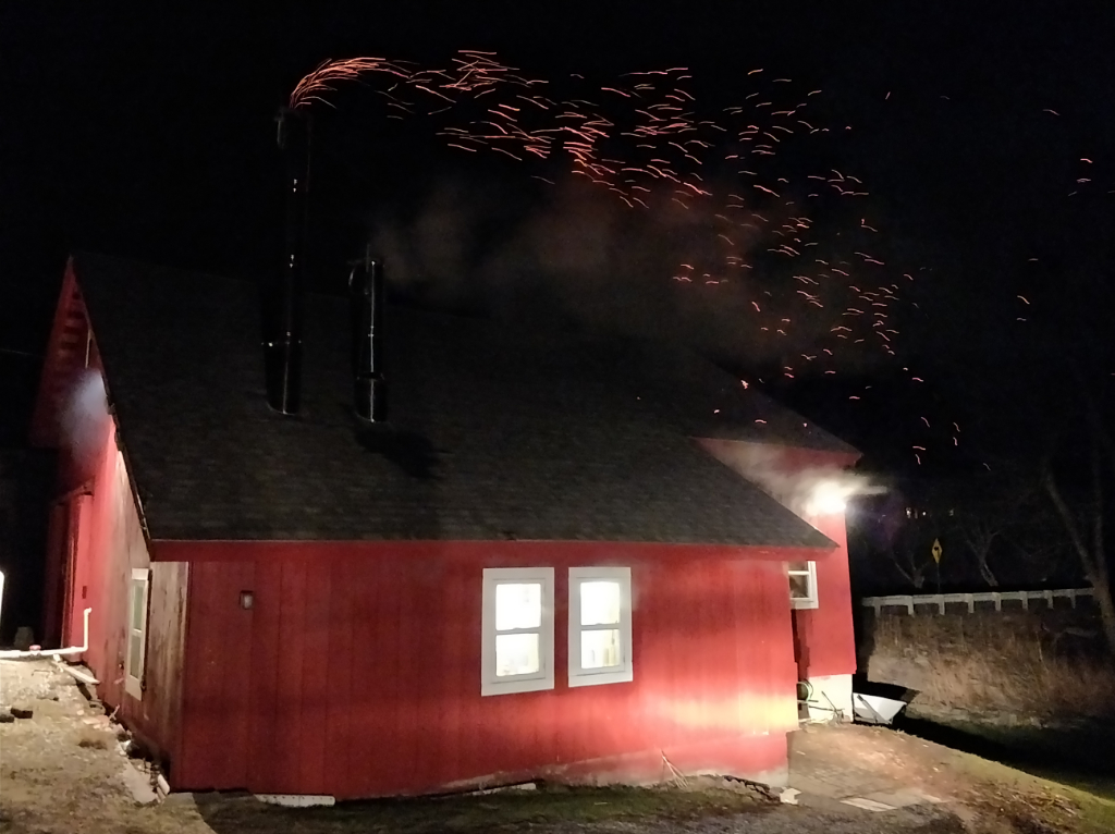 The sugarhouse during a night boil. Steam visible in the windows and embers flying out of the smoke stack.